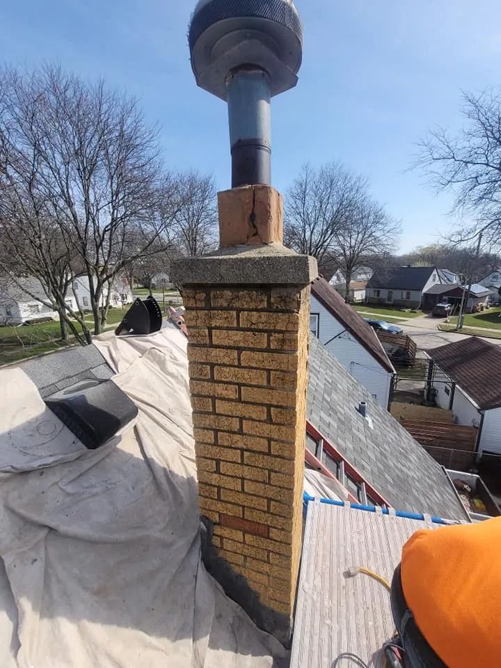 Brick chimney on a roof, topped with a metal flue. Tan tarp covers the roof.