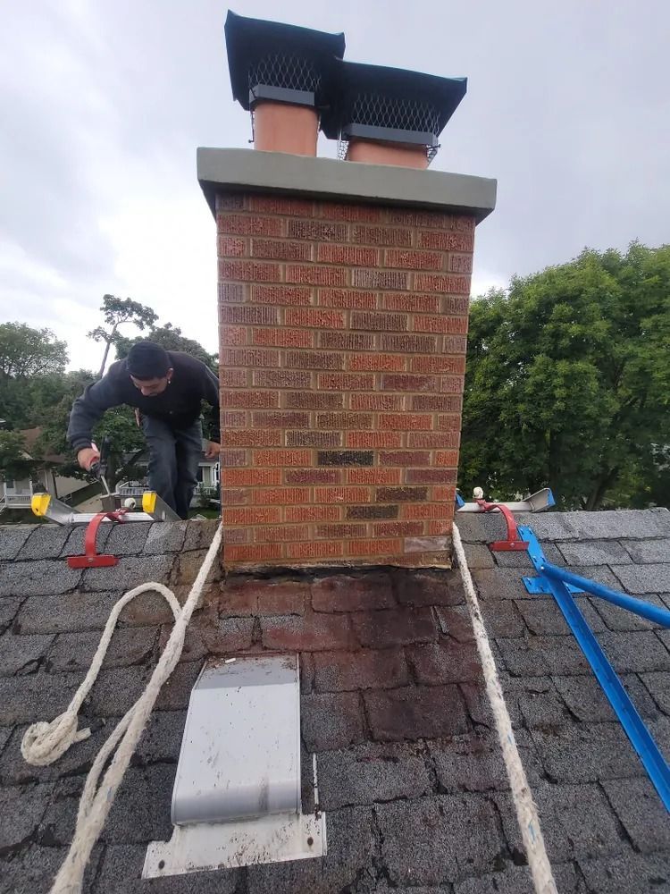 Person on roof repairs brick chimney. Chimney is topped with two metal caps and gray concrete.