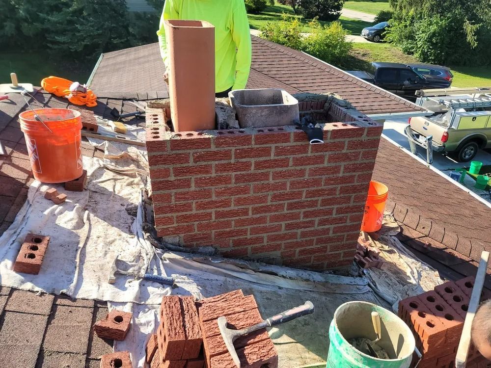 Chimney being rebuilt on a rooftop; bricks, mortar, and a worker in a yellow shirt are visible.