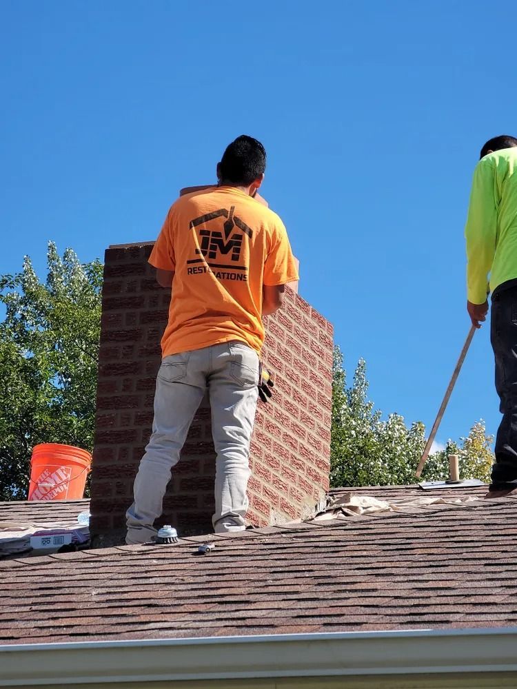 Two workers on a rooftop, one near a brick chimney. Orange shirt, gray pants. Sunny day.