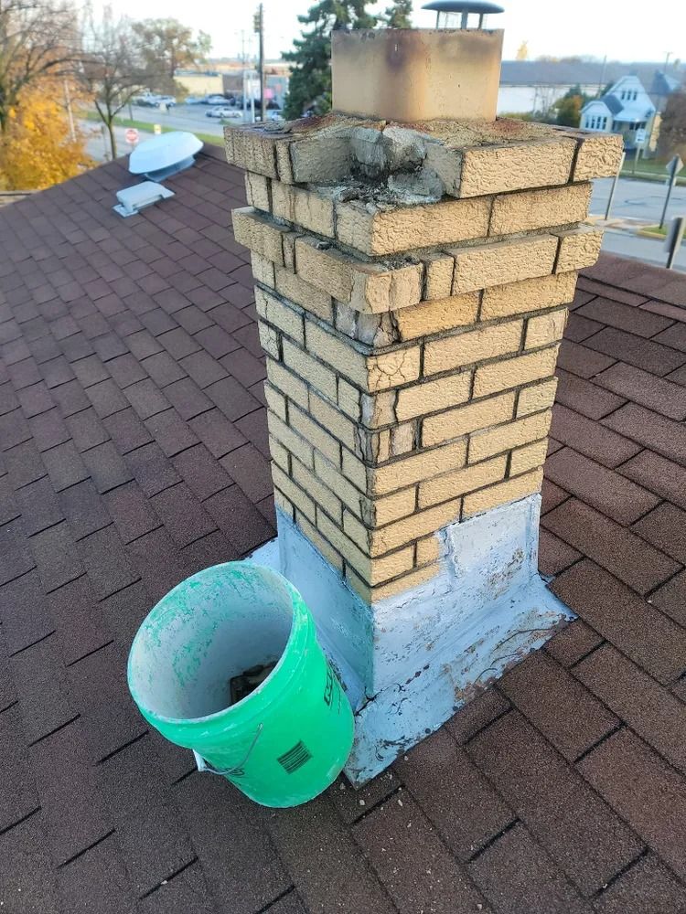A dilapidated brick chimney on a brown shingled roof, next to a green bucket.