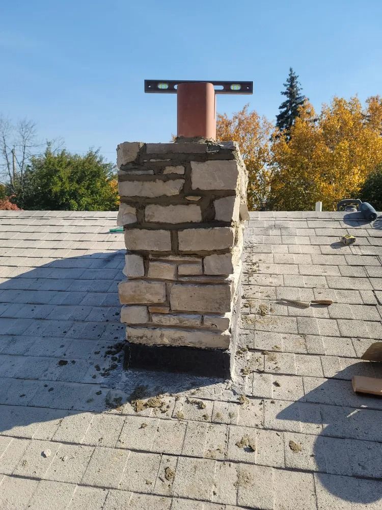 Stone chimney on a shingled roof, with metal bracket and pipe. Blue sky background.