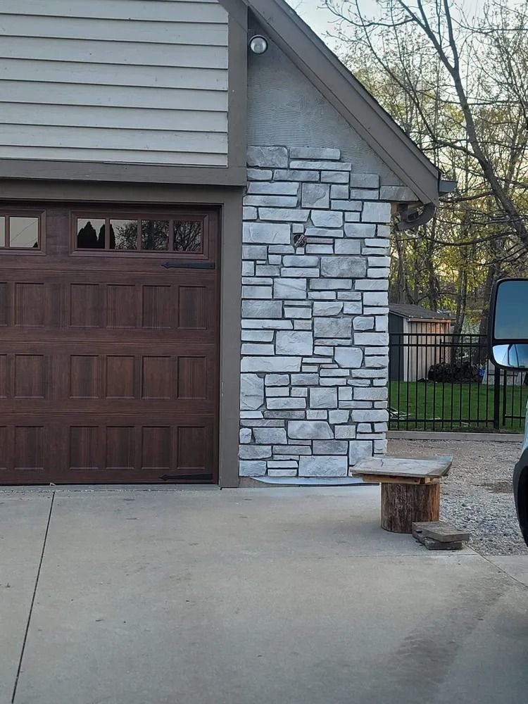 Garage with stone accent, wooden door, and a tree stump bench on a concrete driveway.