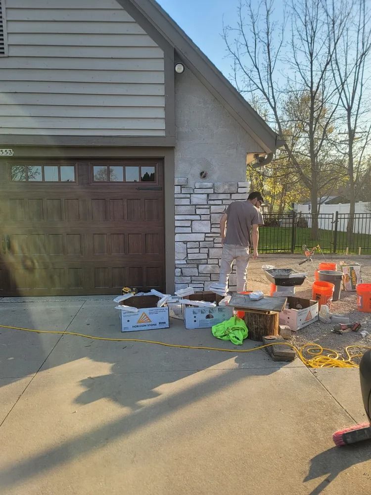 Man installing stone veneer on a house exterior. Tools, materials, and boxes on the driveway.