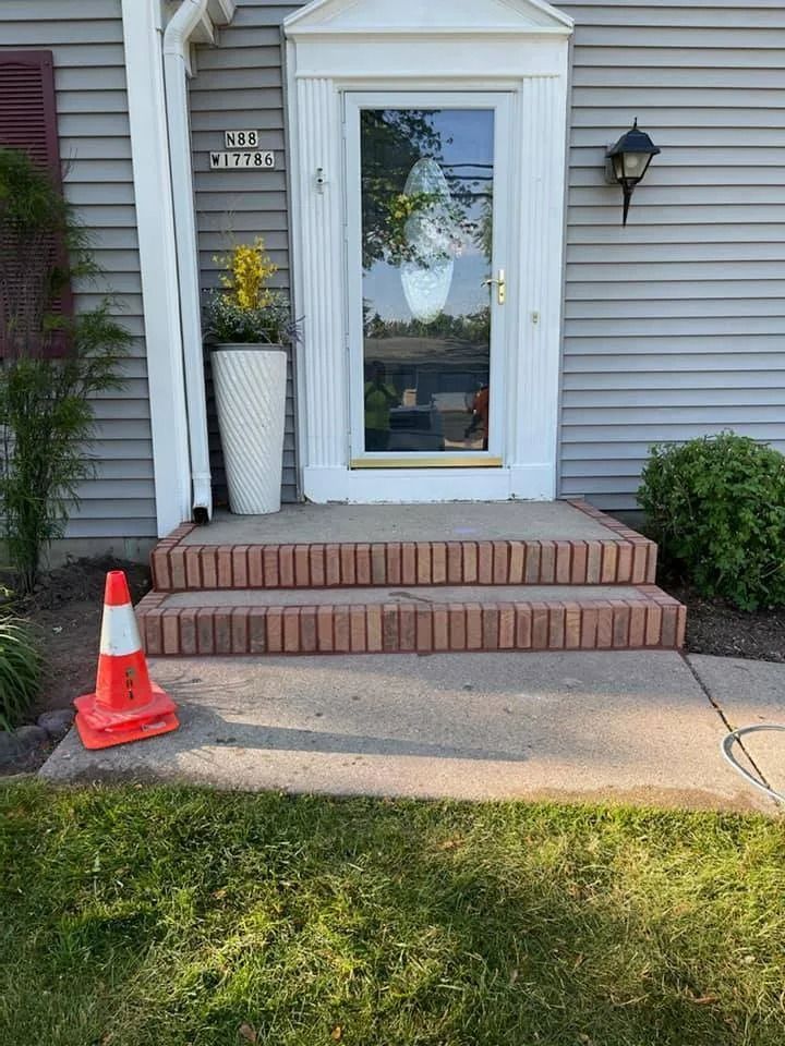 A house entrance with brick steps, a glass door, and a white planter. A traffic cone sits on the lawn.