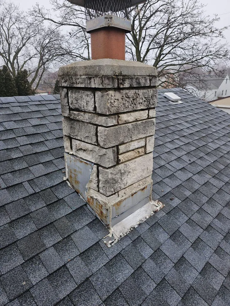 Stone chimney on a gray shingle roof, surrounded by bare trees under a cloudy sky.