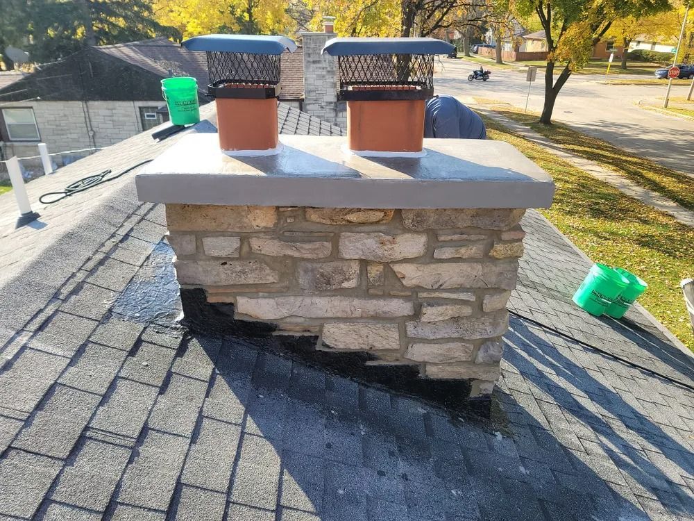 Chimney on a roof; stone construction with two flue caps. Green buckets nearby. Sunny outdoor setting.