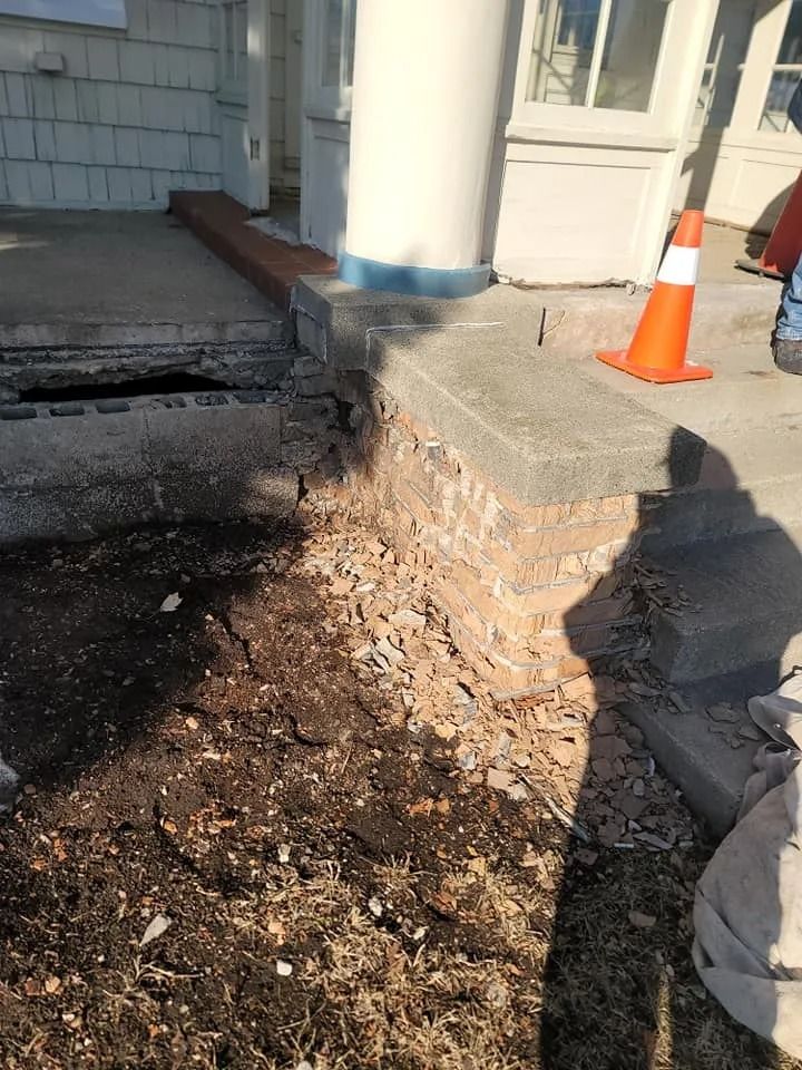 Damaged brick foundation and porch step under repair, next to a white pillar.