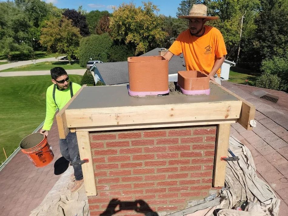 Two workers repairing a brick chimney on a roof, one holding a bucket, the other adjusting a terracotta flue.