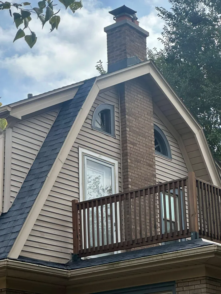 Brick chimney on the roof of a house with light brown siding, a dark roof, and a wooden balcony.