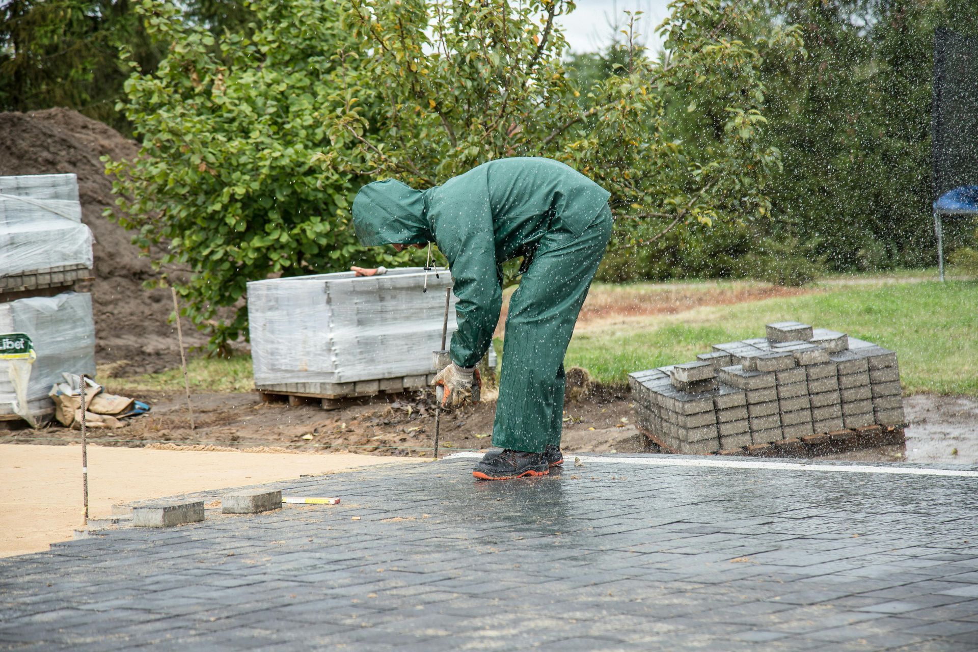 Worker in green coveralls bends over paving stones at an outdoor construction site.