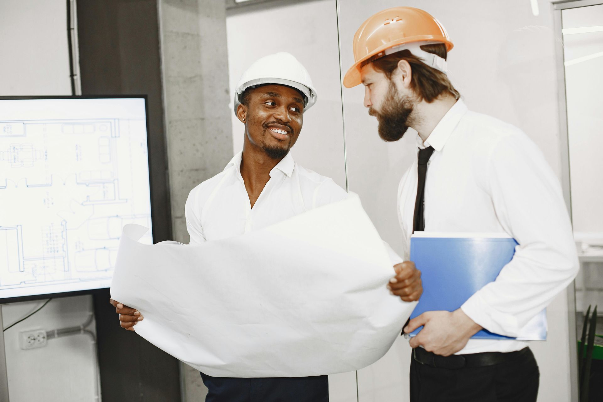 Two professionals in hard hats review a blueprint together in an office setting.