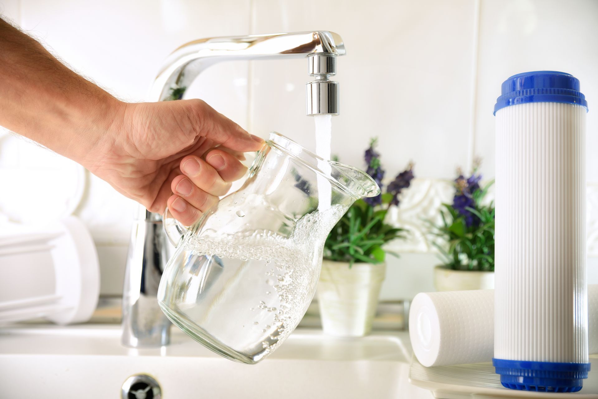 Hand pouring water from a glass pitcher into a kitchen sink with a faucet and water filter nearby