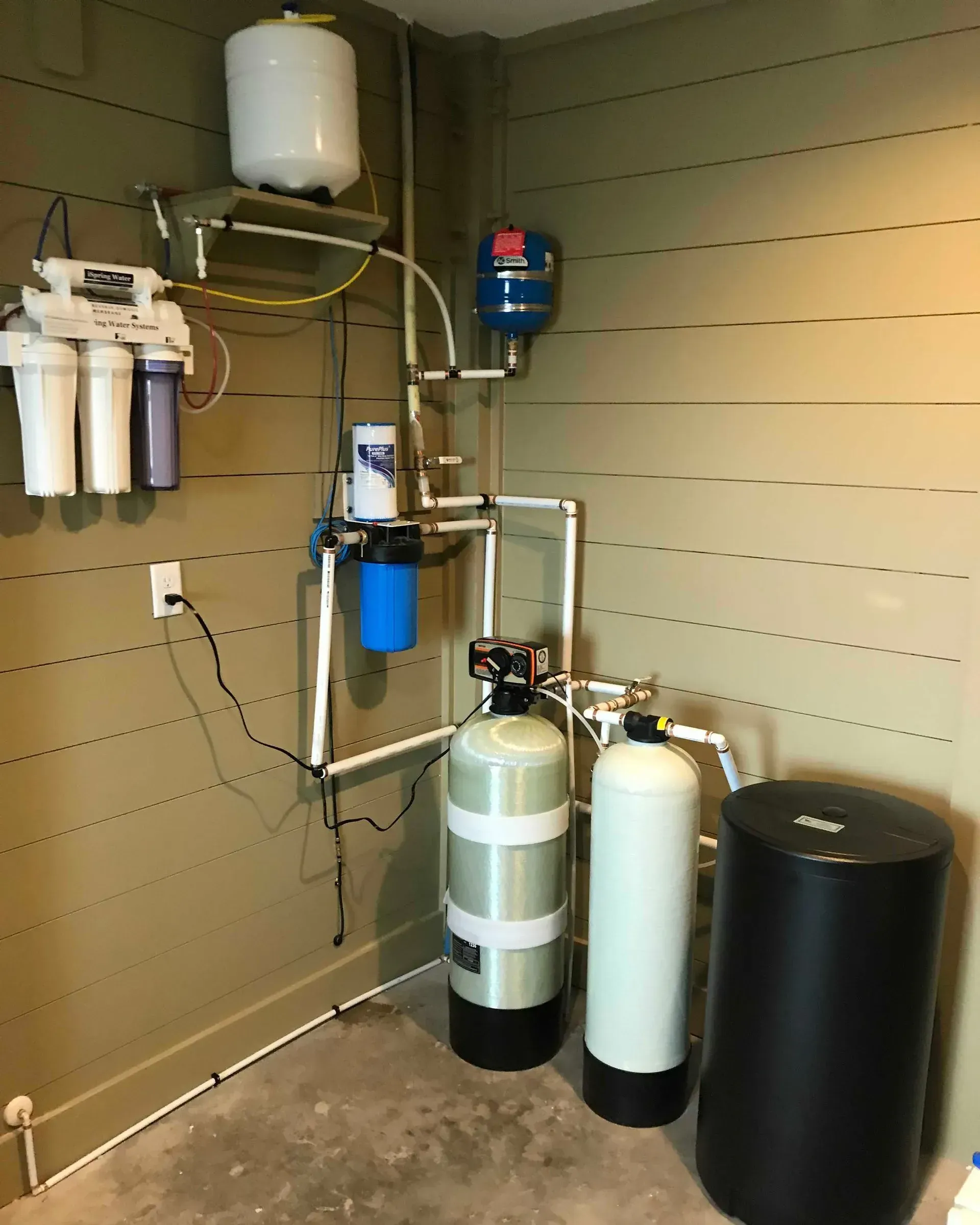 Water filtration equipment in a utility room, with tanks, canisters, pipes, and wall-mounted filters.