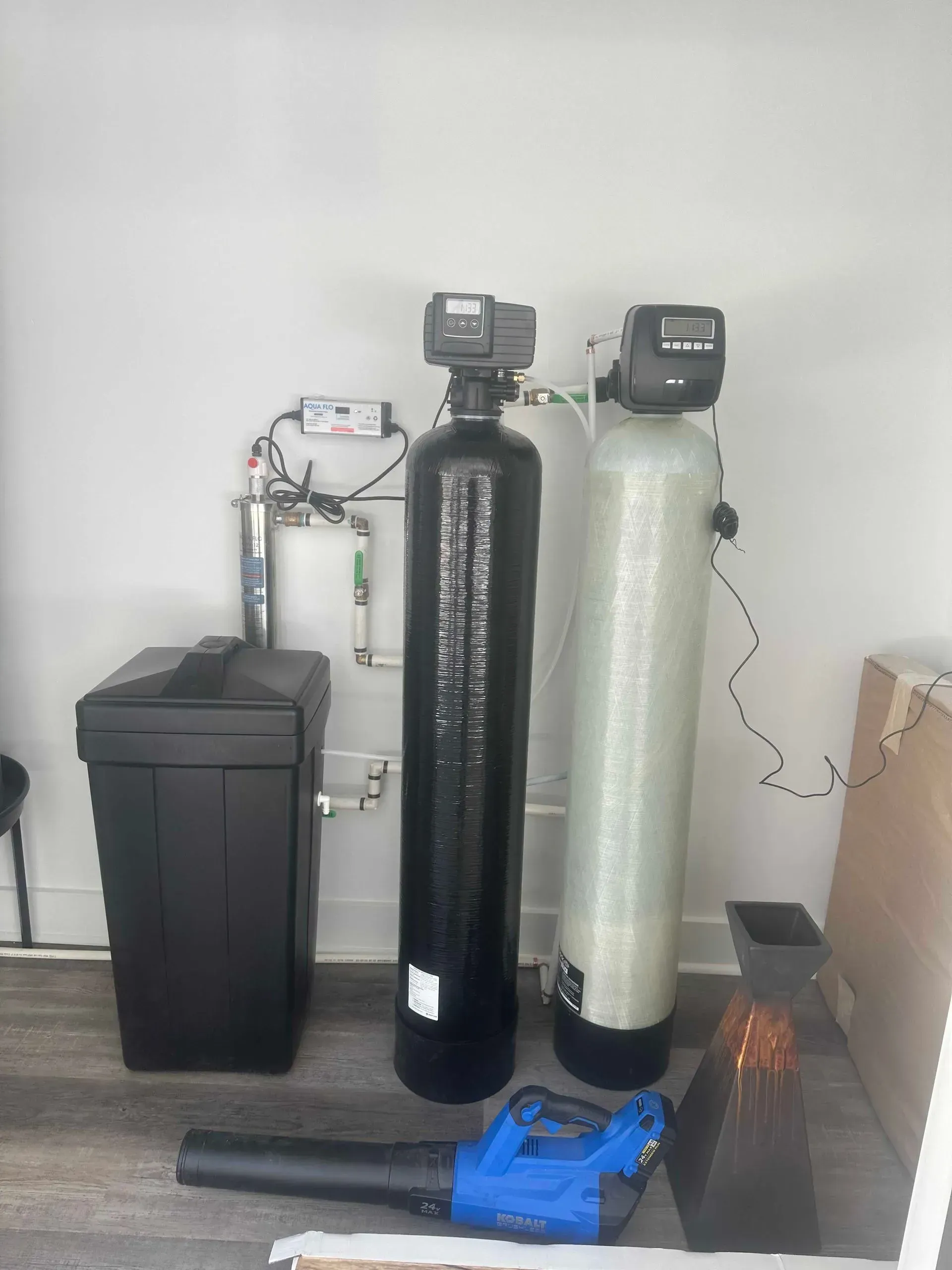 Water softener and filtration system with black and white tanks, blue tools, and a black bin in a utility room