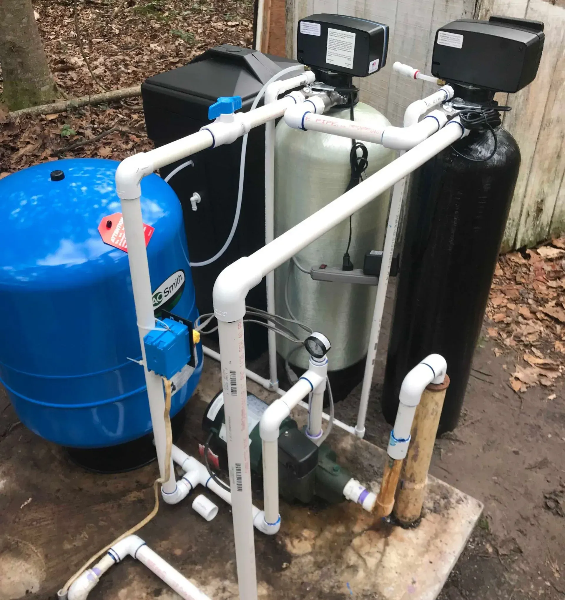 Outdoor water filtration setup with blue and black tanks, white PVC pipes, and pump on a concrete pad
