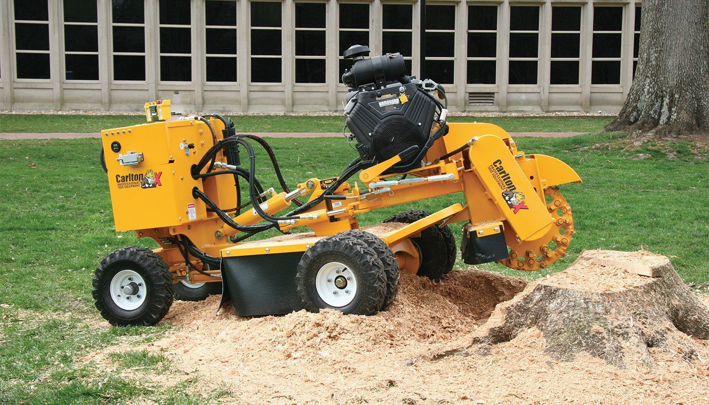 A yellow stump grinder is cutting a tree stump in the grass.