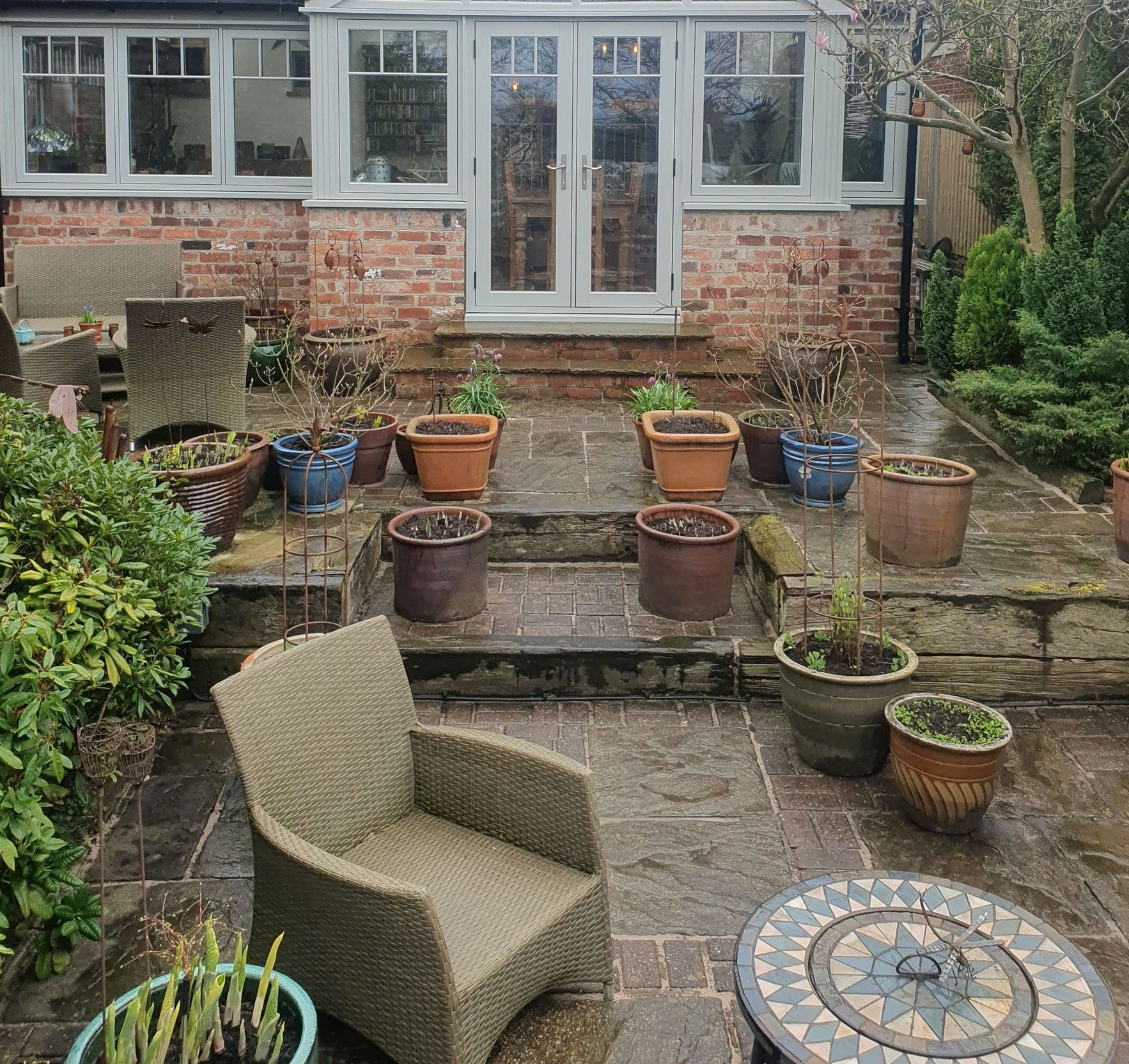 Backyard patio with steps leading to a sunroom with potted plants, brick wall, and a wicker chair.