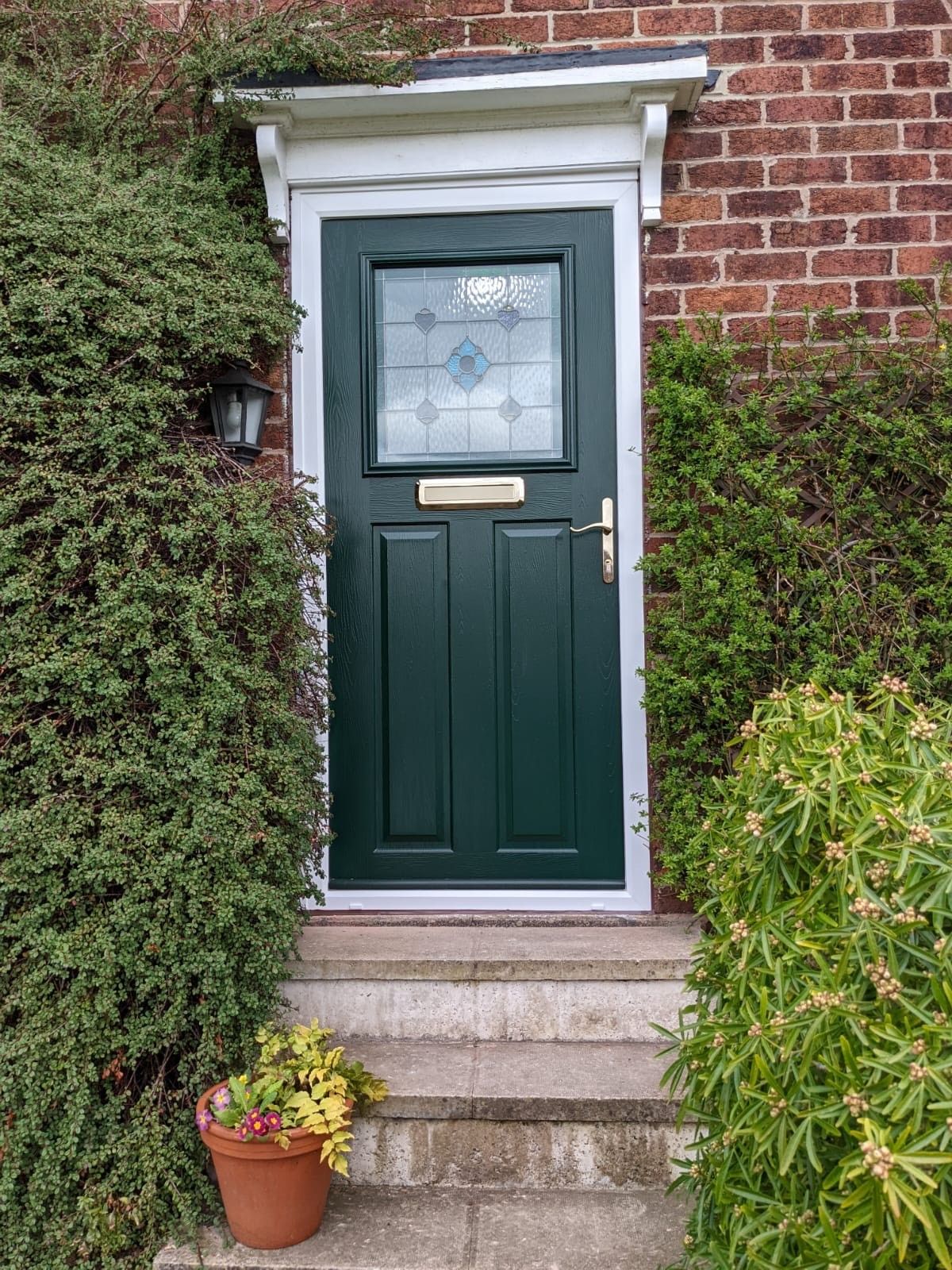 Green front door with glass panel, white trim, and a small porch roof, framed by brick and greenery.