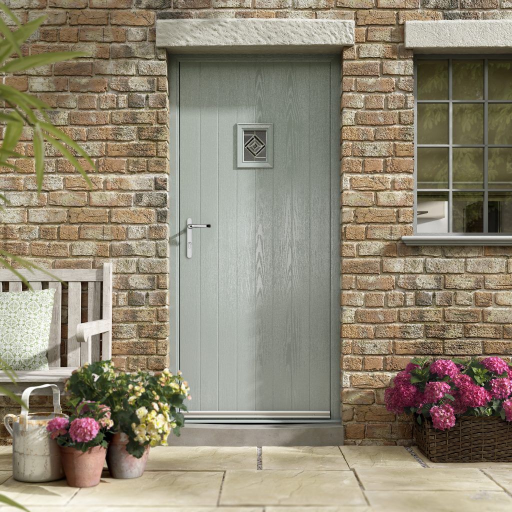 Gray-green front door with small window, set in a brick wall. Flowers and a bench decorate the porch.