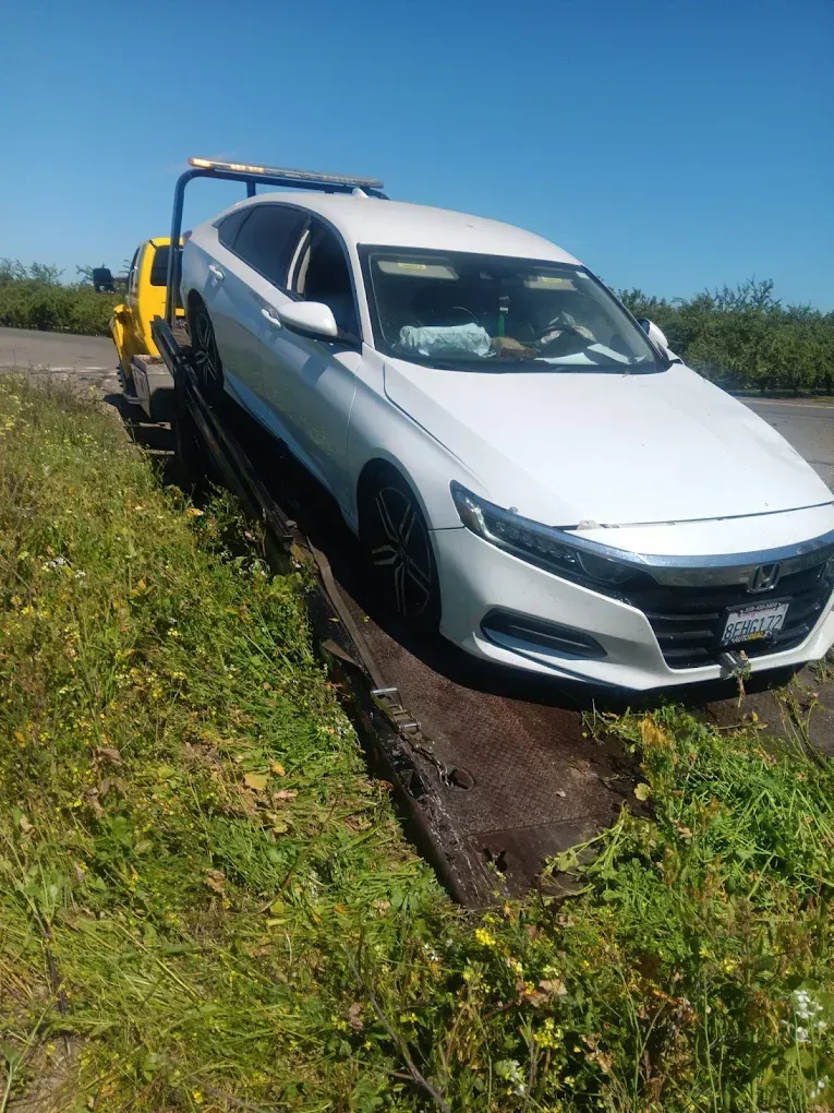 A white Honda sedan being loaded onto the flatbed of a yellow tow truck on a roadside with grass.