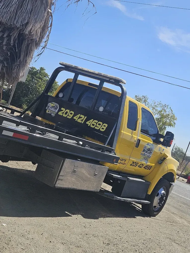 A bright yellow flatbed work truck parked on a gravel lot under a clear sky, showing its phone number on the side.