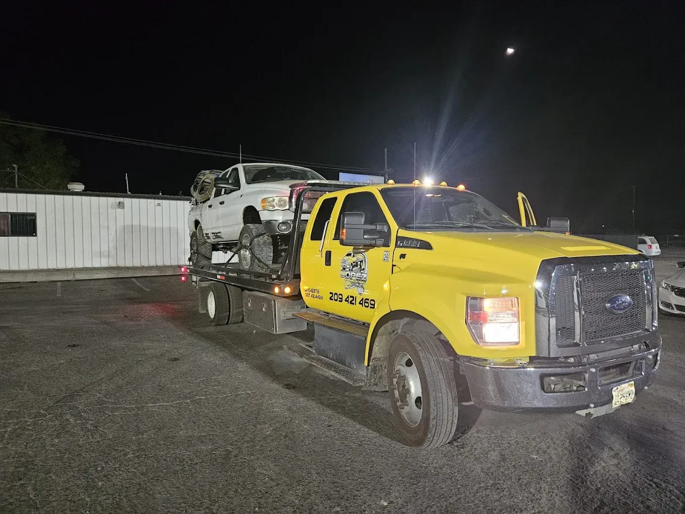 A bright yellow flatbed tow truck parked at night, carrying a white pickup truck on its bed.