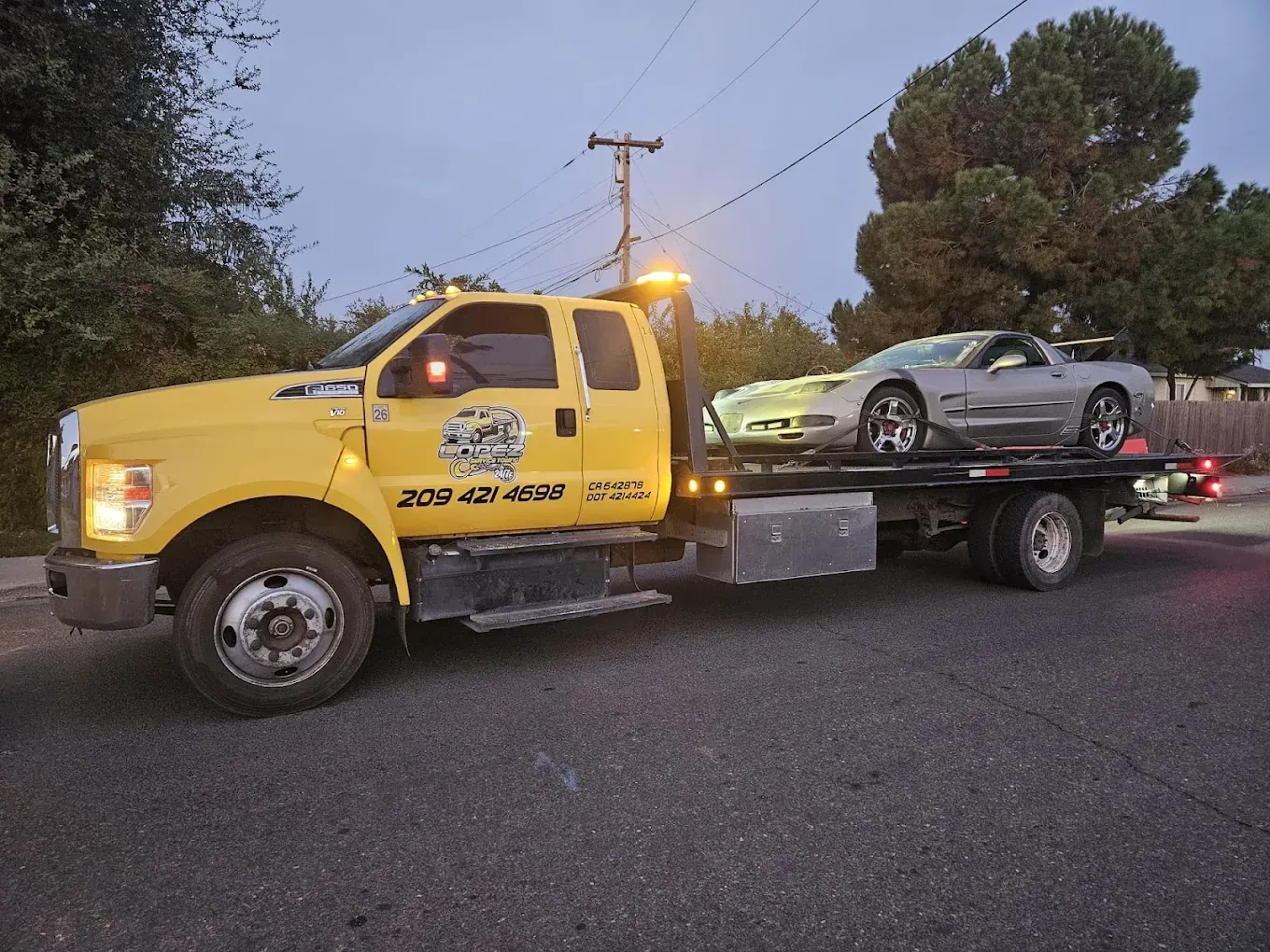 A yellow flatbed tow truck carrying a silver sports car on a residential street during twilight.