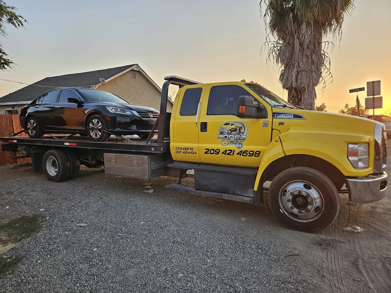 A yellow flatbed tow truck carrying a black sedan parked on a gravel lot during sunset.