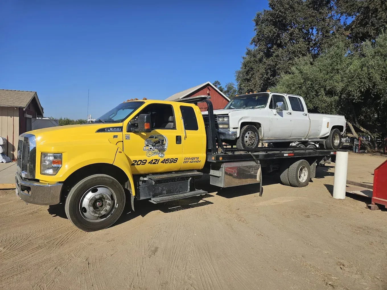 A yellow tow truck parked on a dirt lot carrying a white pickup truck on its flatbed.