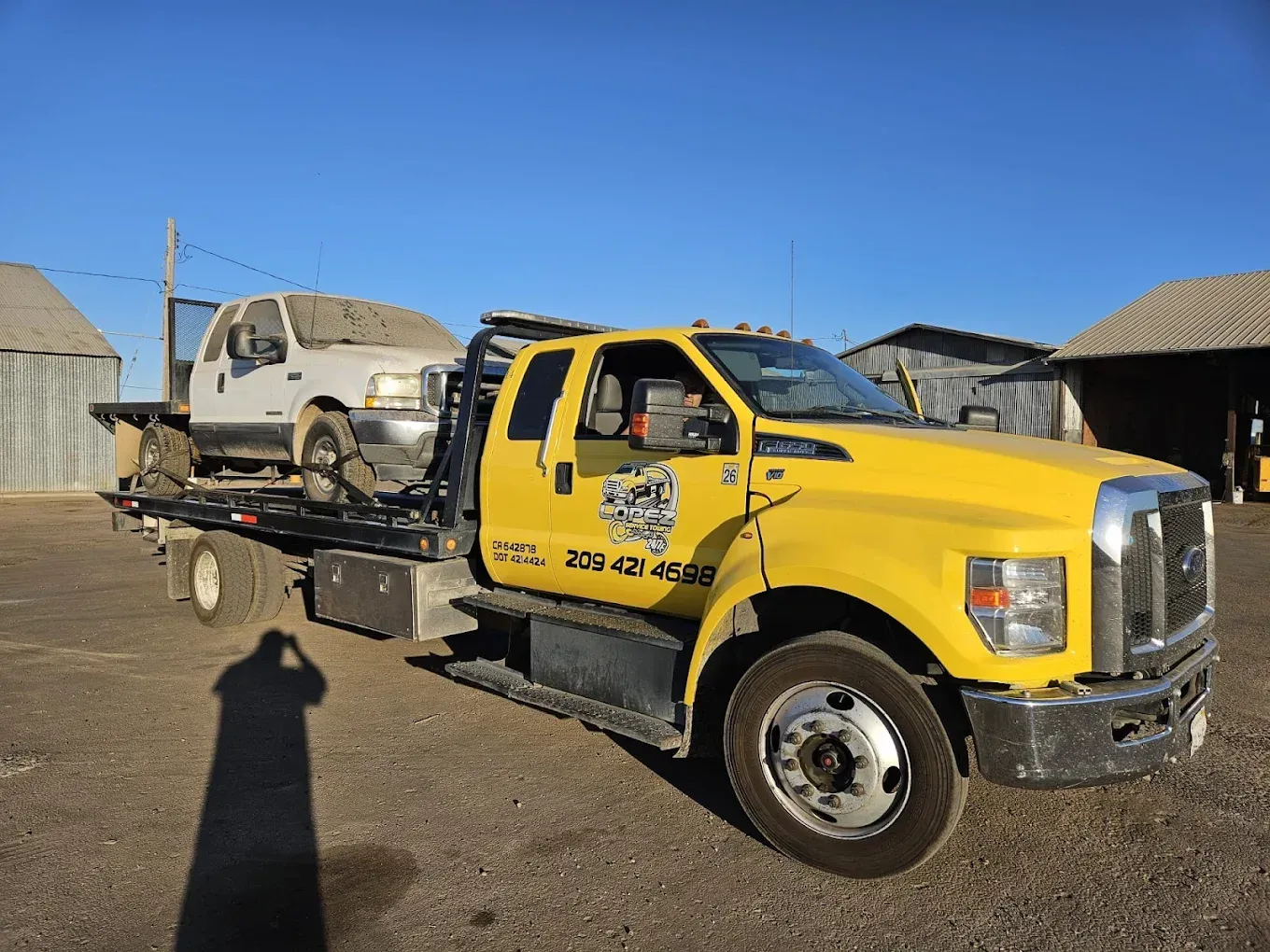 A bright yellow flatbed tow truck parked outdoors carrying a white pickup truck, with a building visible in the background.