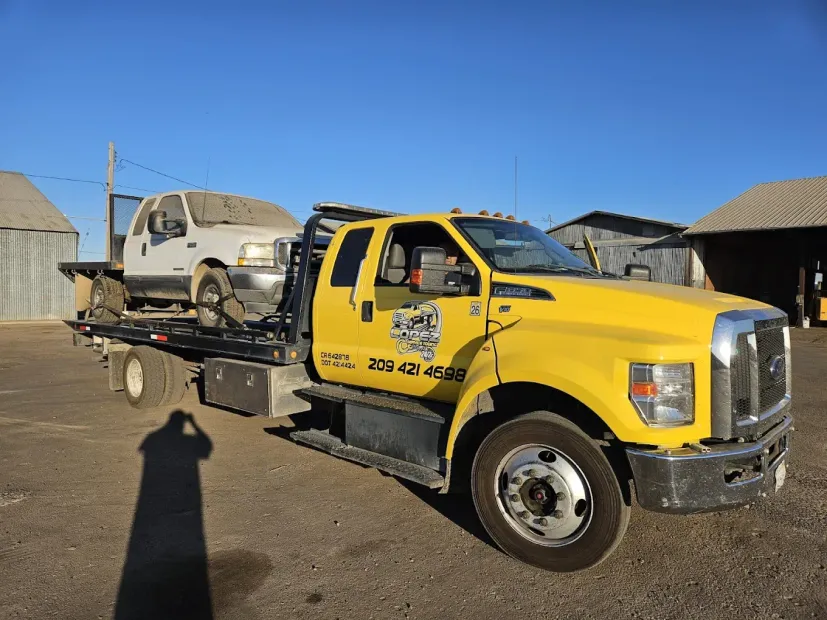 A bright yellow flatbed tow truck parked outdoors carrying a white pickup truck, with a building visible in the background.