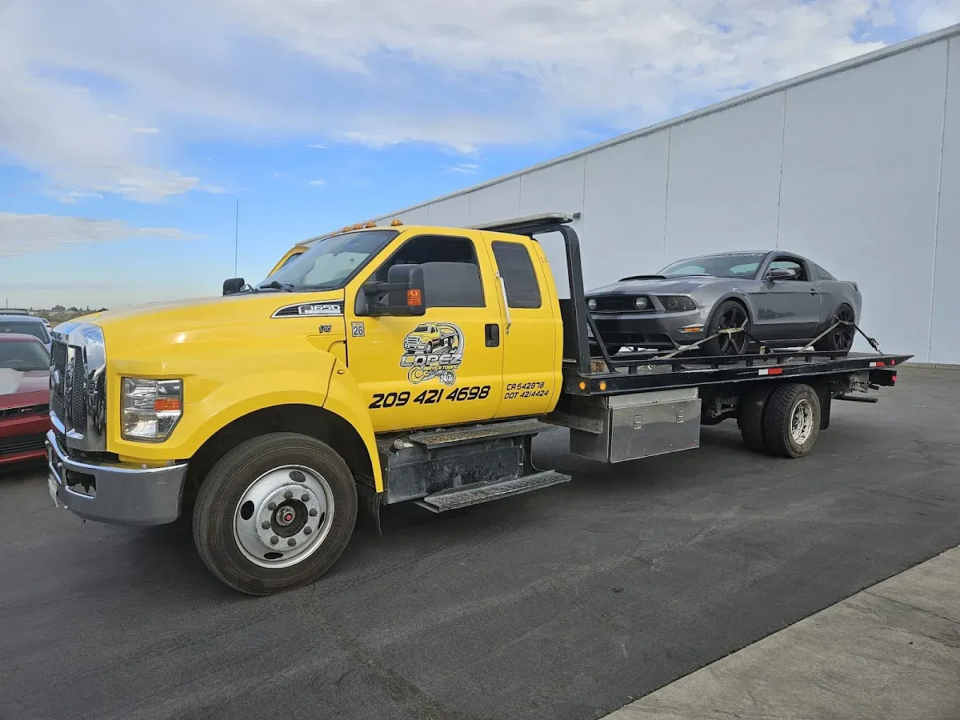 A yellow flatbed tow truck carrying a gray Ford Mustang in an open parking lot against a large white building.