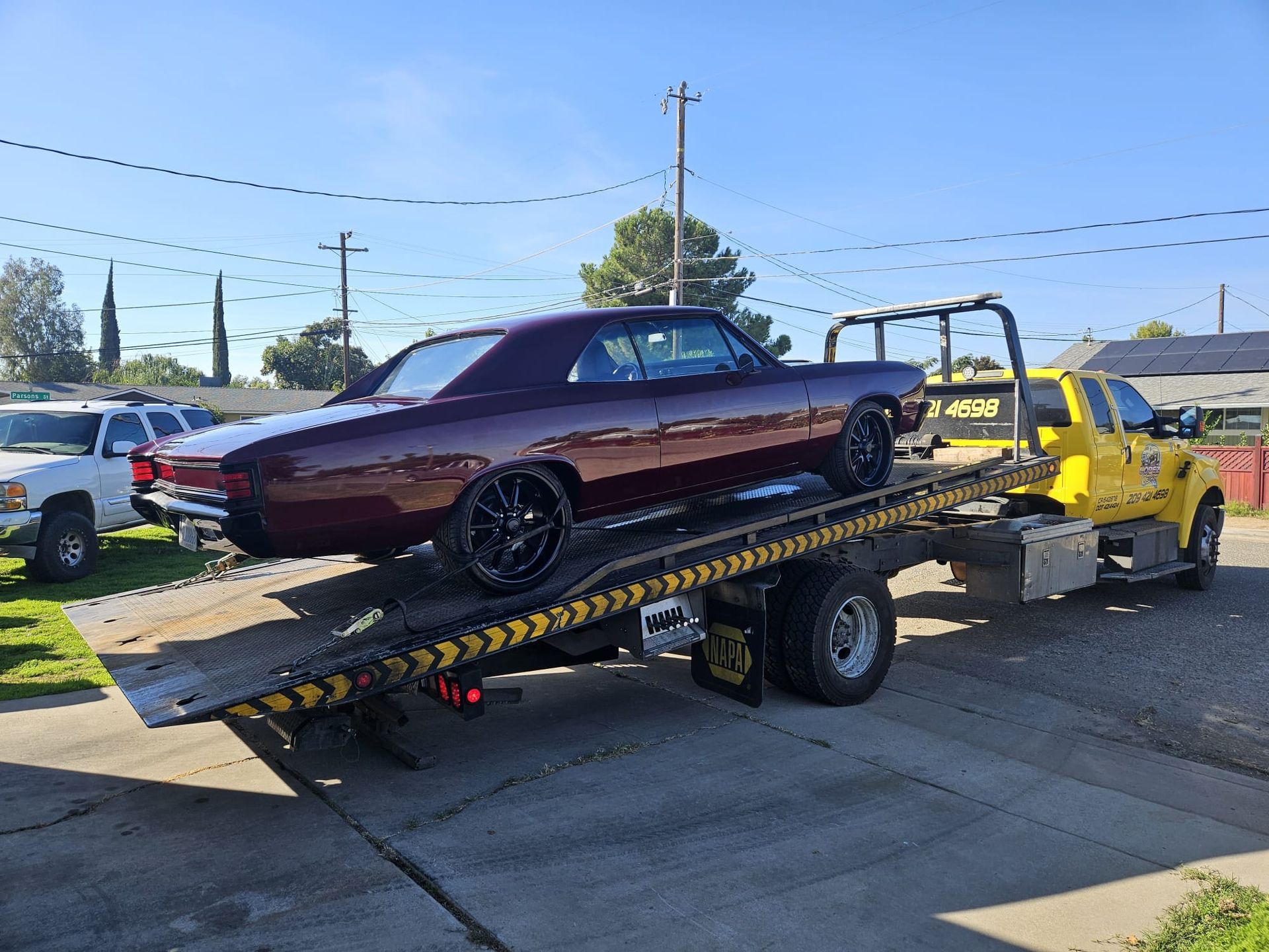 A dark purple vintage muscle car loaded onto a yellow flatbed tow truck on a sunny day.