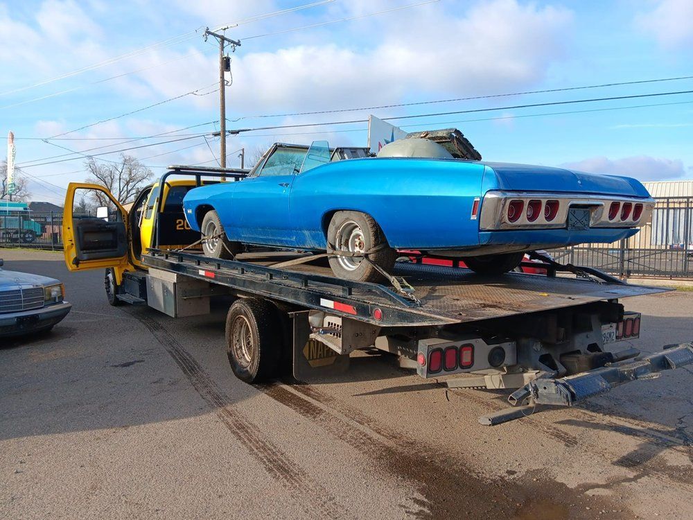 A bright blue classic convertible car secured on the flatbed of a yellow tow truck in an outdoor parking lot.