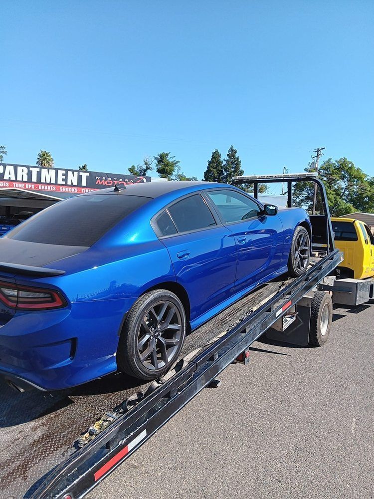 A bright blue Dodge Charger loaded onto a black flatbed tow truck on a sunny day.
