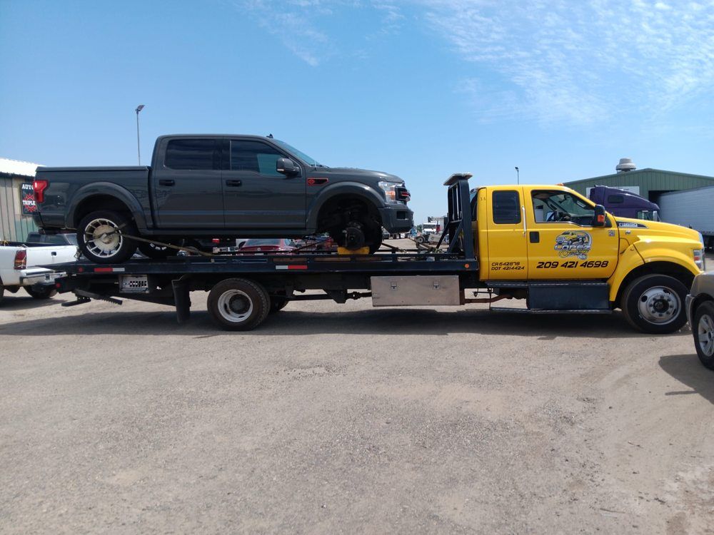 A yellow flatbed tow truck transporting a gray pickup truck that is missing its front passenger-side wheel.