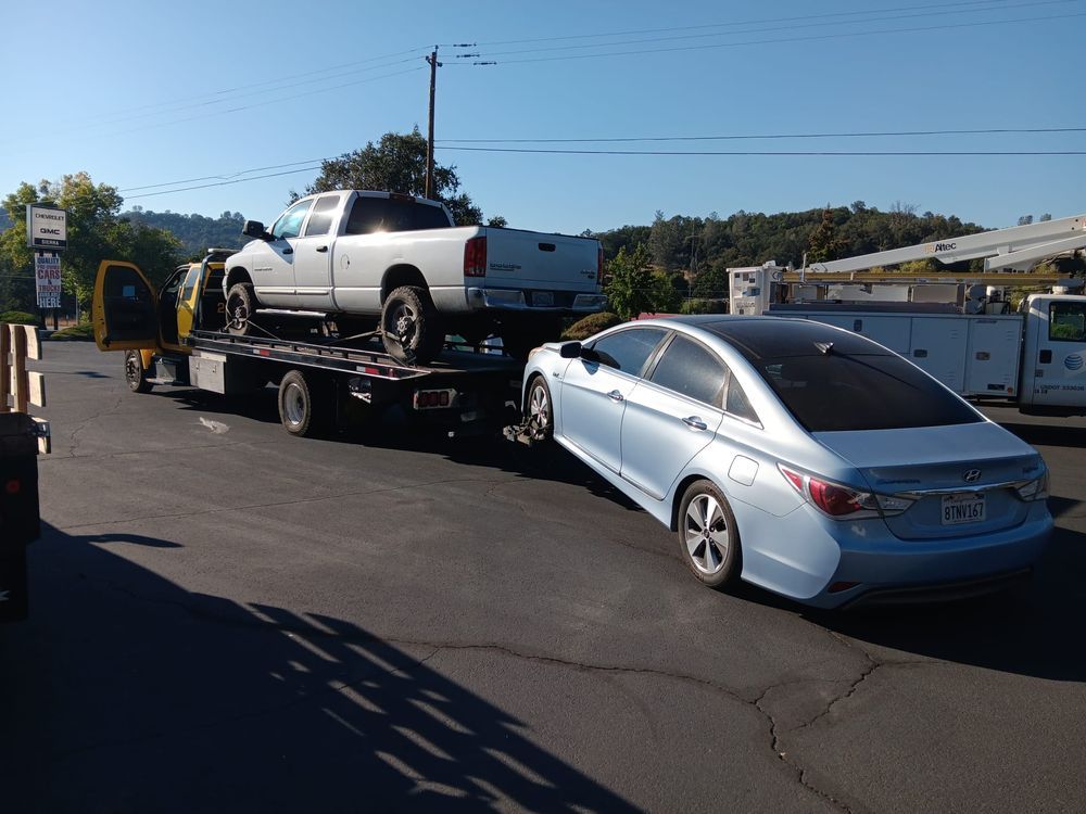 A white pickup truck sits on a tow truck flatbed while a light blue sedan is hitched to the rear for towing.