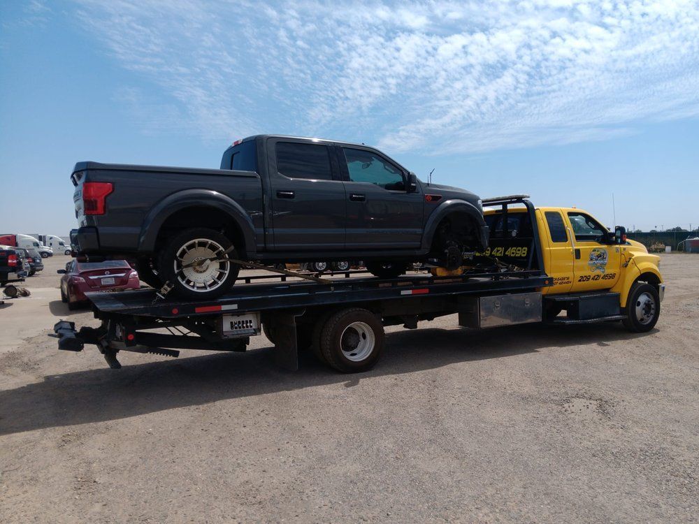A grey pickup truck is being transported on the flatbed of a yellow tow truck in a gravel lot under a sunny sky.