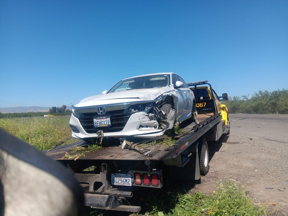 A damaged white Honda sedan loaded onto a yellow flatbed tow truck on a rural roadside under a clear blue sky.