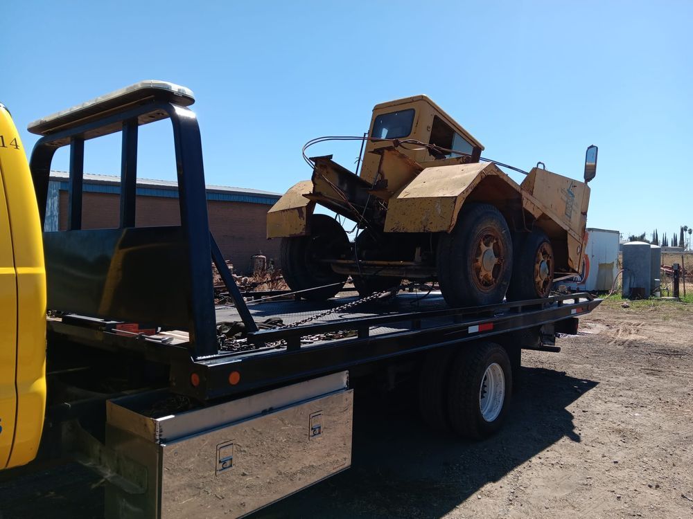 A yellow, dismantled industrial loader sits strapped to the flatbed of a yellow tow truck on a sunny day.