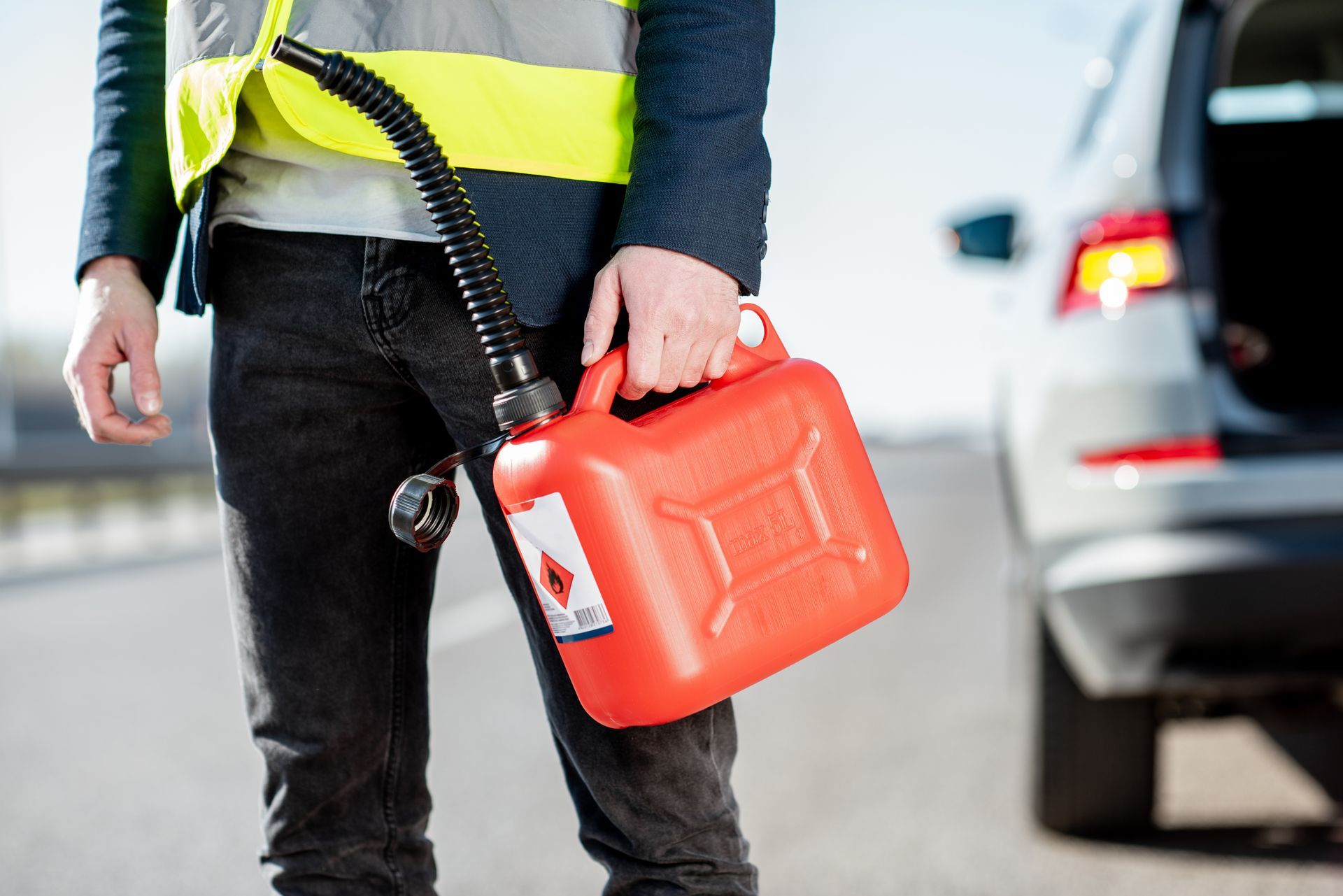 A person in a reflective safety vest holds a red fuel can by the side of a road near a car.