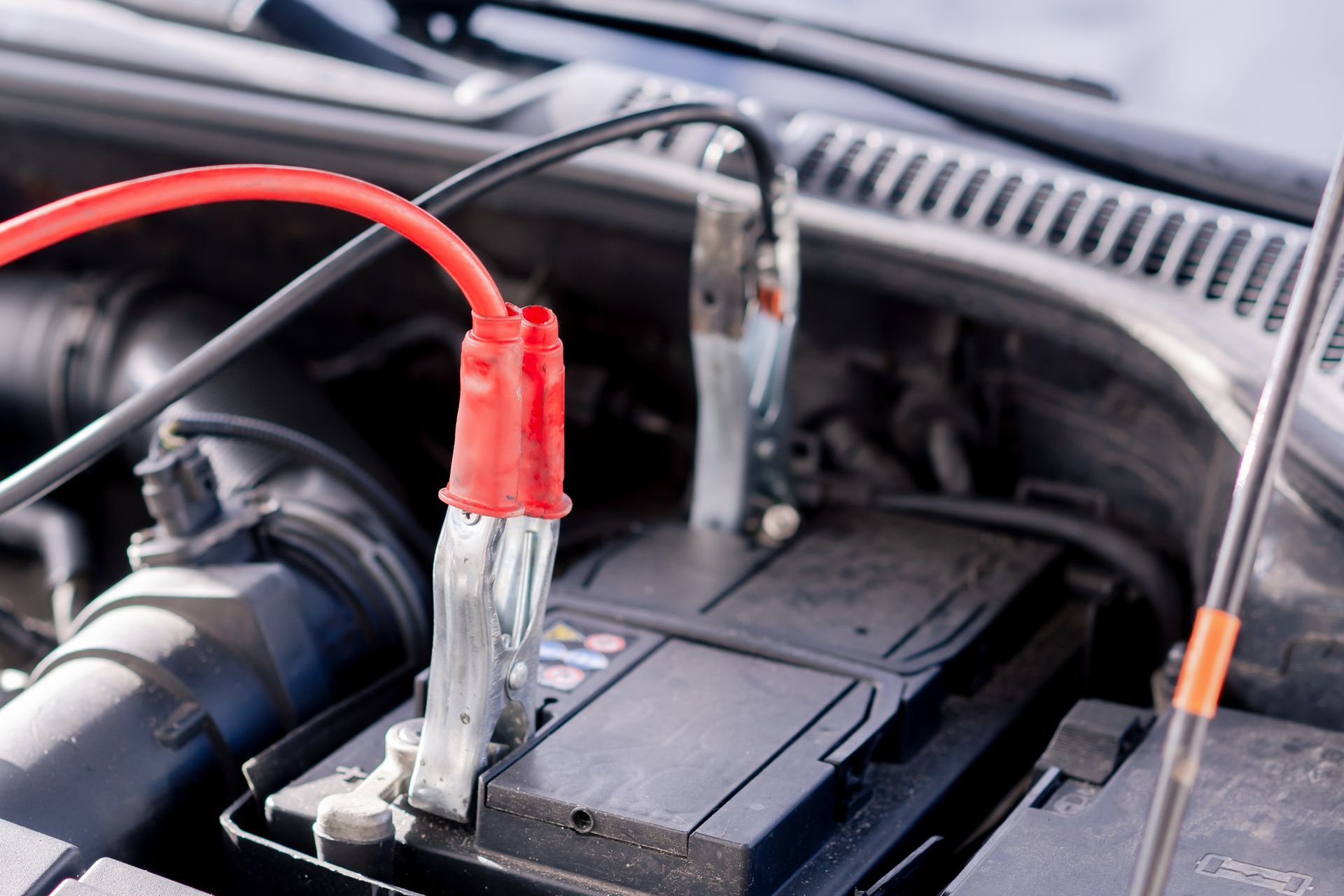 A car battery under a hood with jumper cables attached to the terminals.