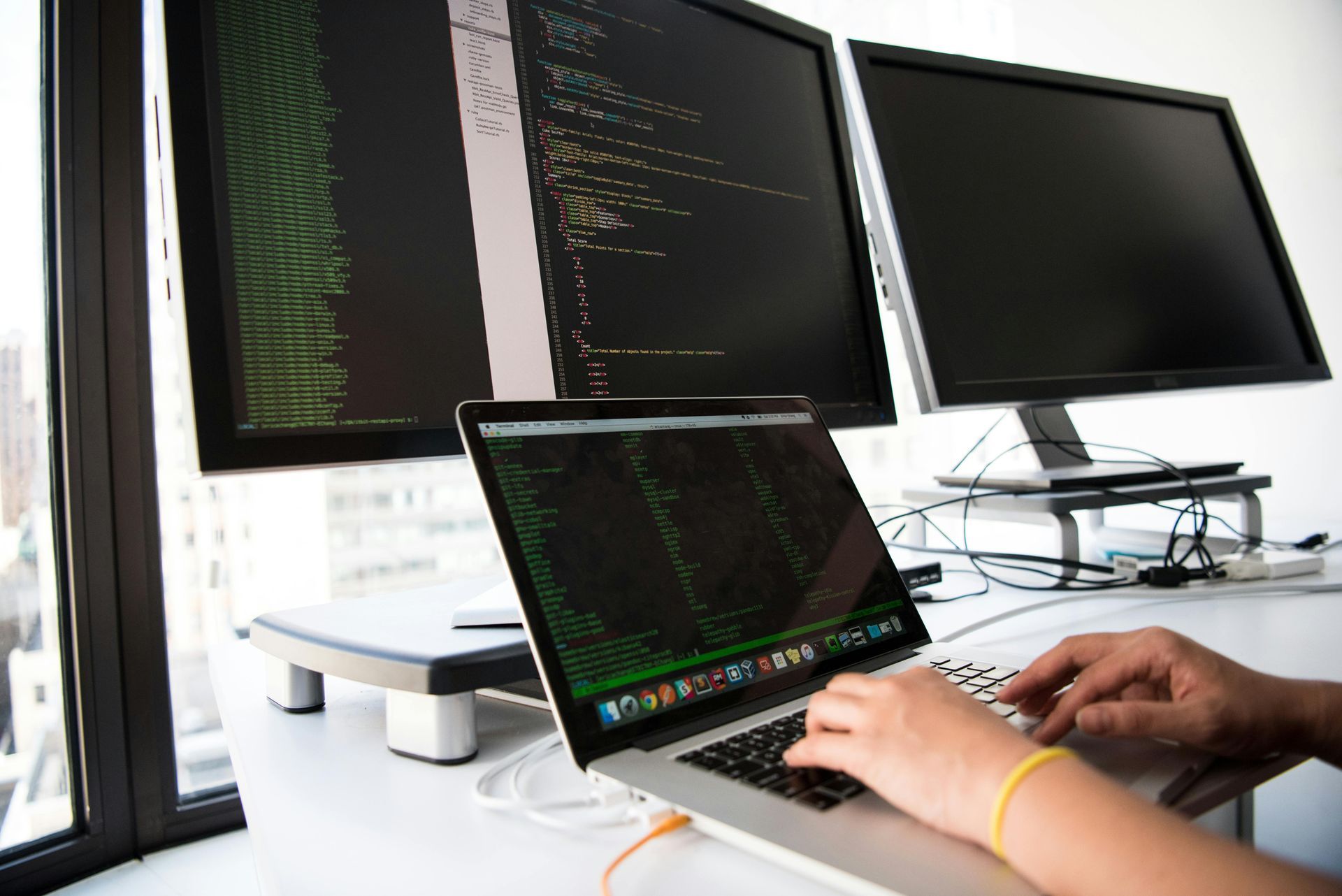 Person typing on a laptop beside dual monitors displaying code in a bright workspace