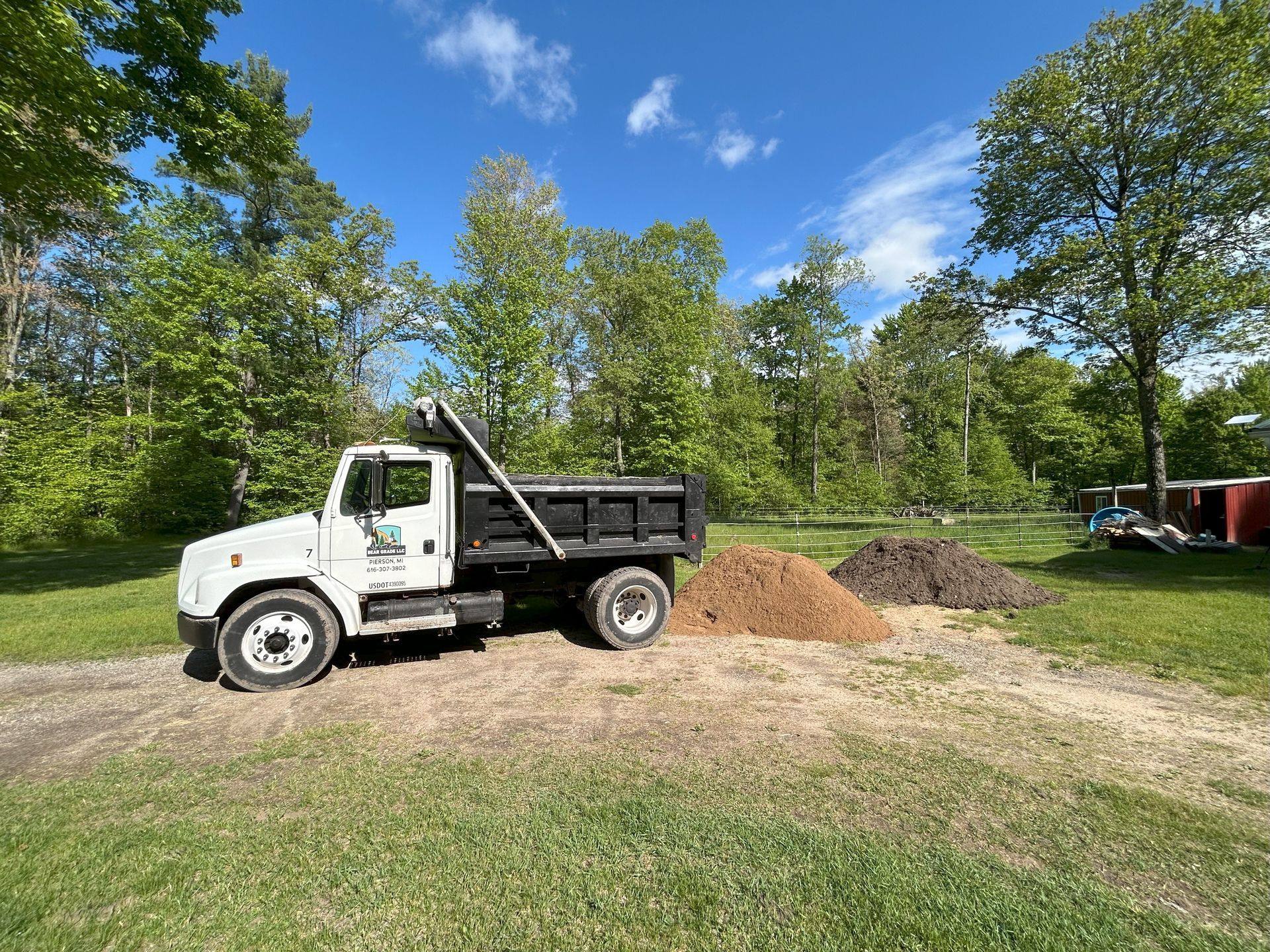 White dump truck unloading soil piles on a grassy area, trees in background under a blue sky.