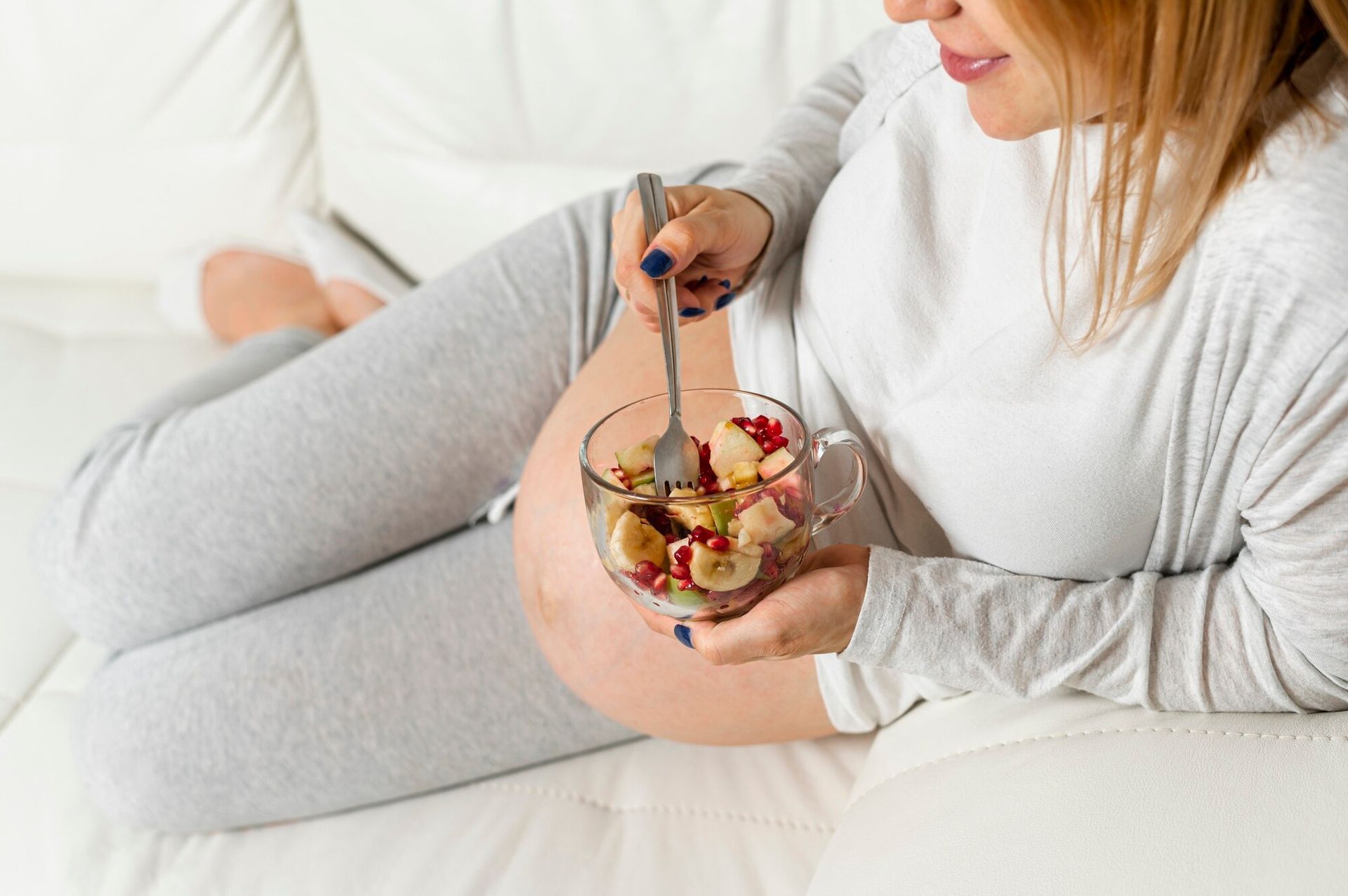 A pregnant woman comfortably sits on the couch and eats a healthy, fruity snack. 