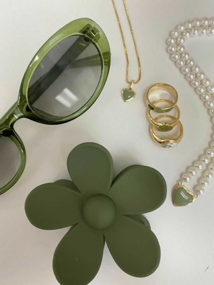 Um par de óculos de sol verdes, uma flor verde, um colar de pérolas e anéis estão sobre uma mesa.