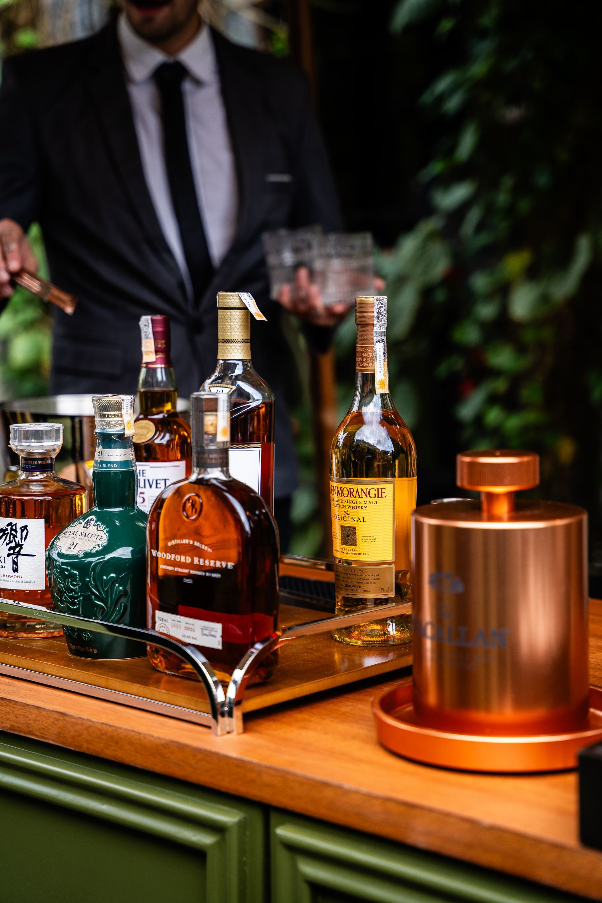 Um homem de terno e gravata está parado atrás de um bar cheio de garrafas de álcool.