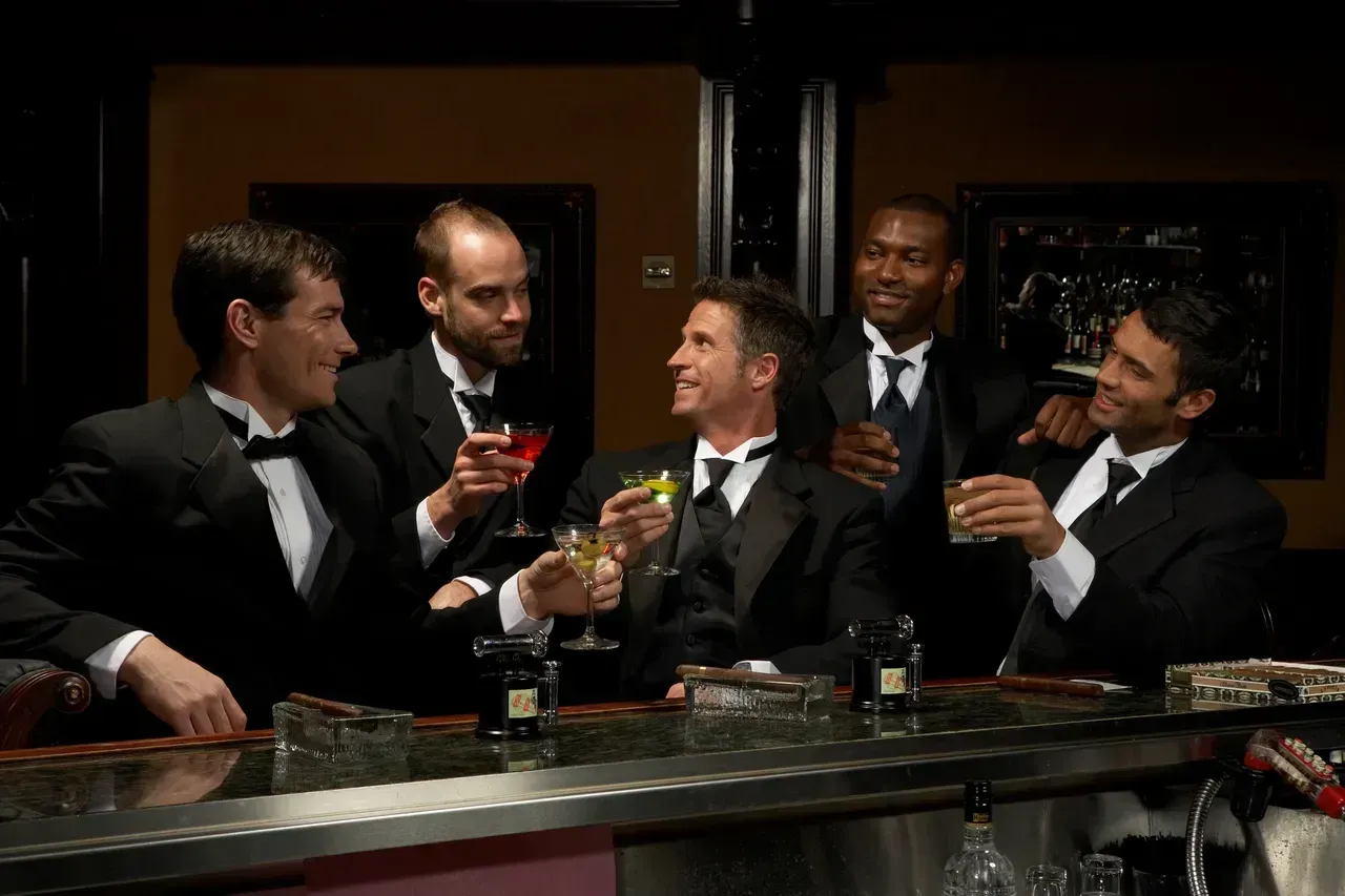 Five men in tuxedos at a bar, toasting with cocktails. Dark interior with a bar.