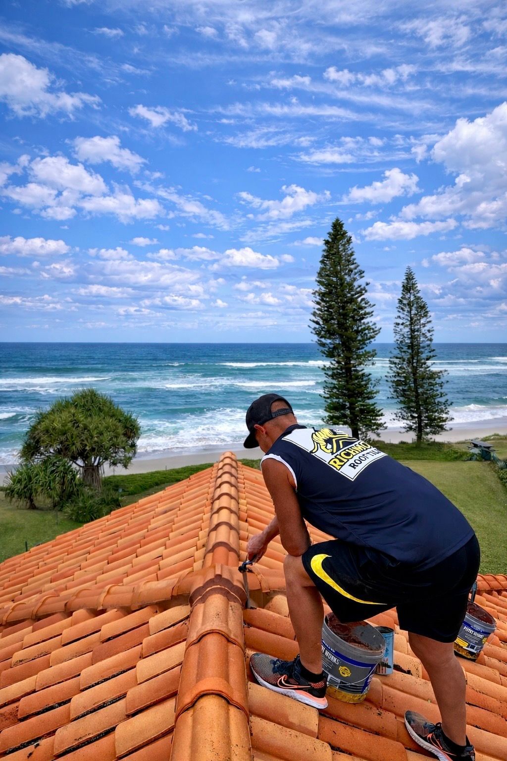 Person on a terracotta tile roof, working with tools. Ocean and beach view with blue sky— Richmond Roof Tiling in Ballina, NSW
